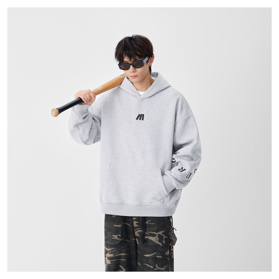 Model wearing a light gray oversized hoodie with a black chest mark and sleeve lettering; he holds a wooden bat.