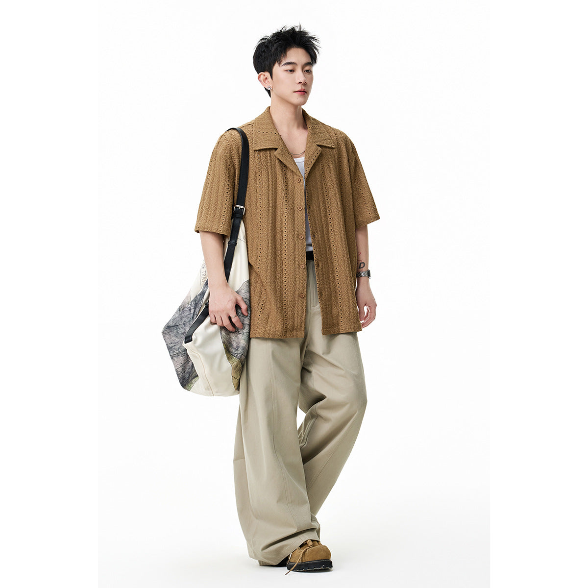 Model wearing a brown short-sleeve eyelet shirt and loose beige trousers, carrying a tote against a white background.