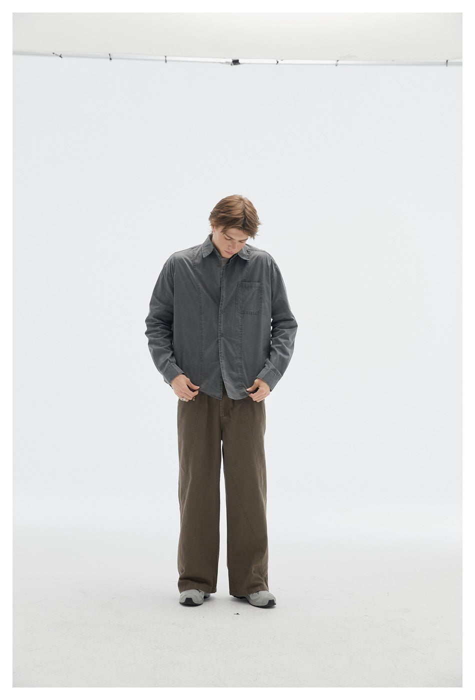 Model wears a buttoned charcoal shirt with a chest pocket and dark olive wide-leg trousers, shown against a plain studio