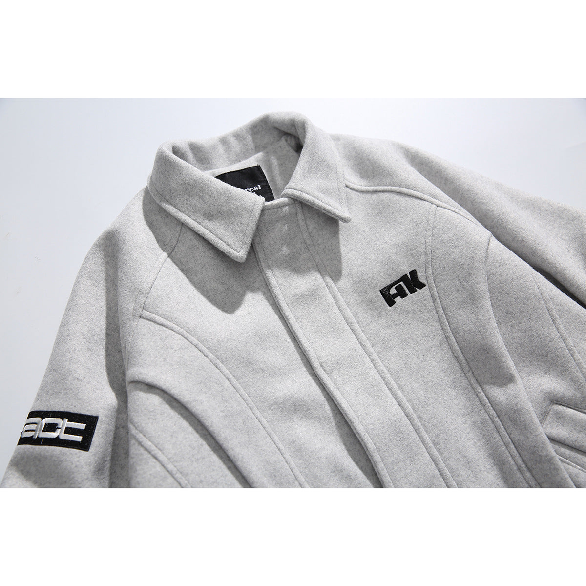 Close-up of a light gray jacket showing its collar, front closure, curved seams, and black embroidered patches.