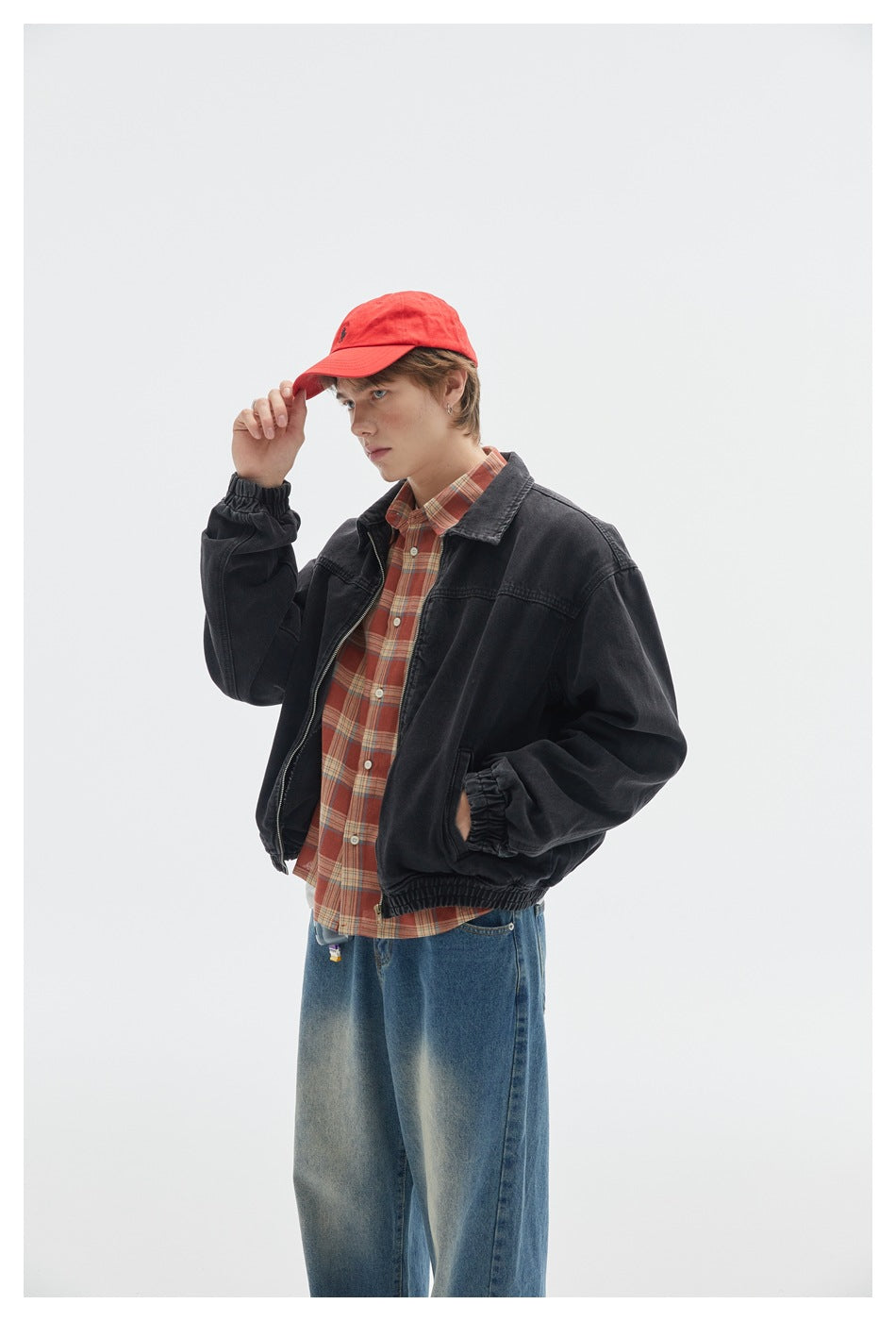 Closer view of a model wearing a black jacket over a red plaid shirt and blue jeans, with a red cap.