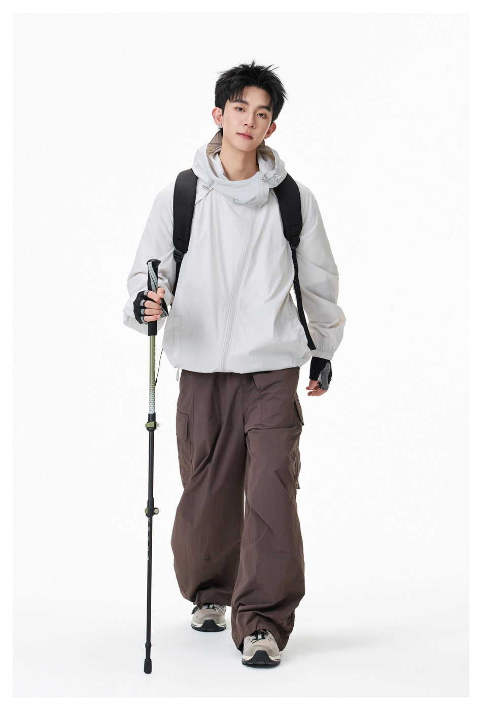 Model walking in a light gray-white hooded jacket with backpack, brown cargo pants and trekking pole