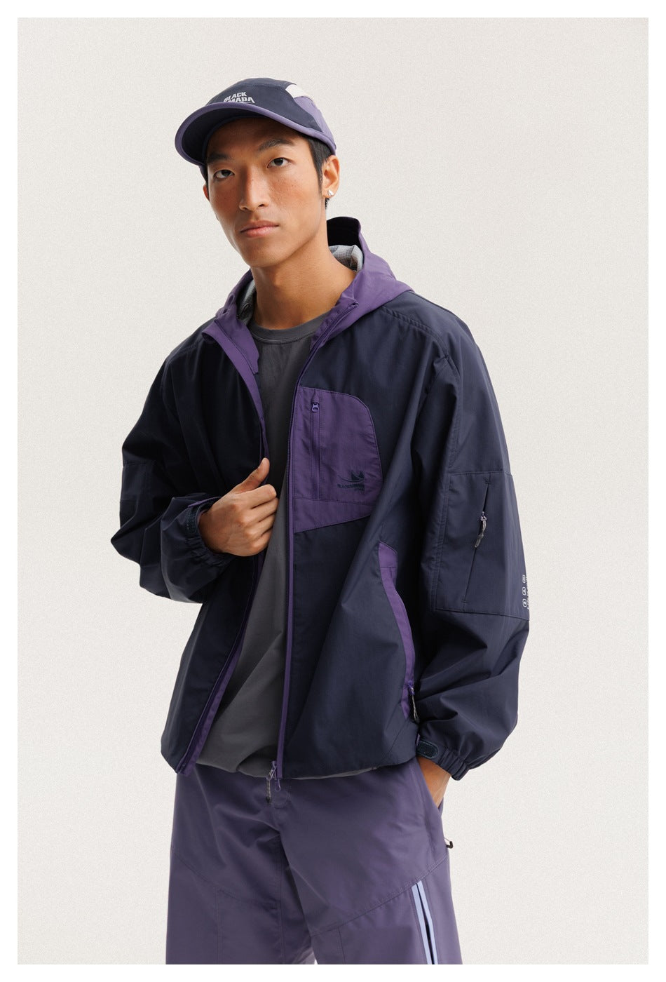 Three-quarter view of a model wearing an open navy jacket with purple hood and chest pocket, and purple pants.