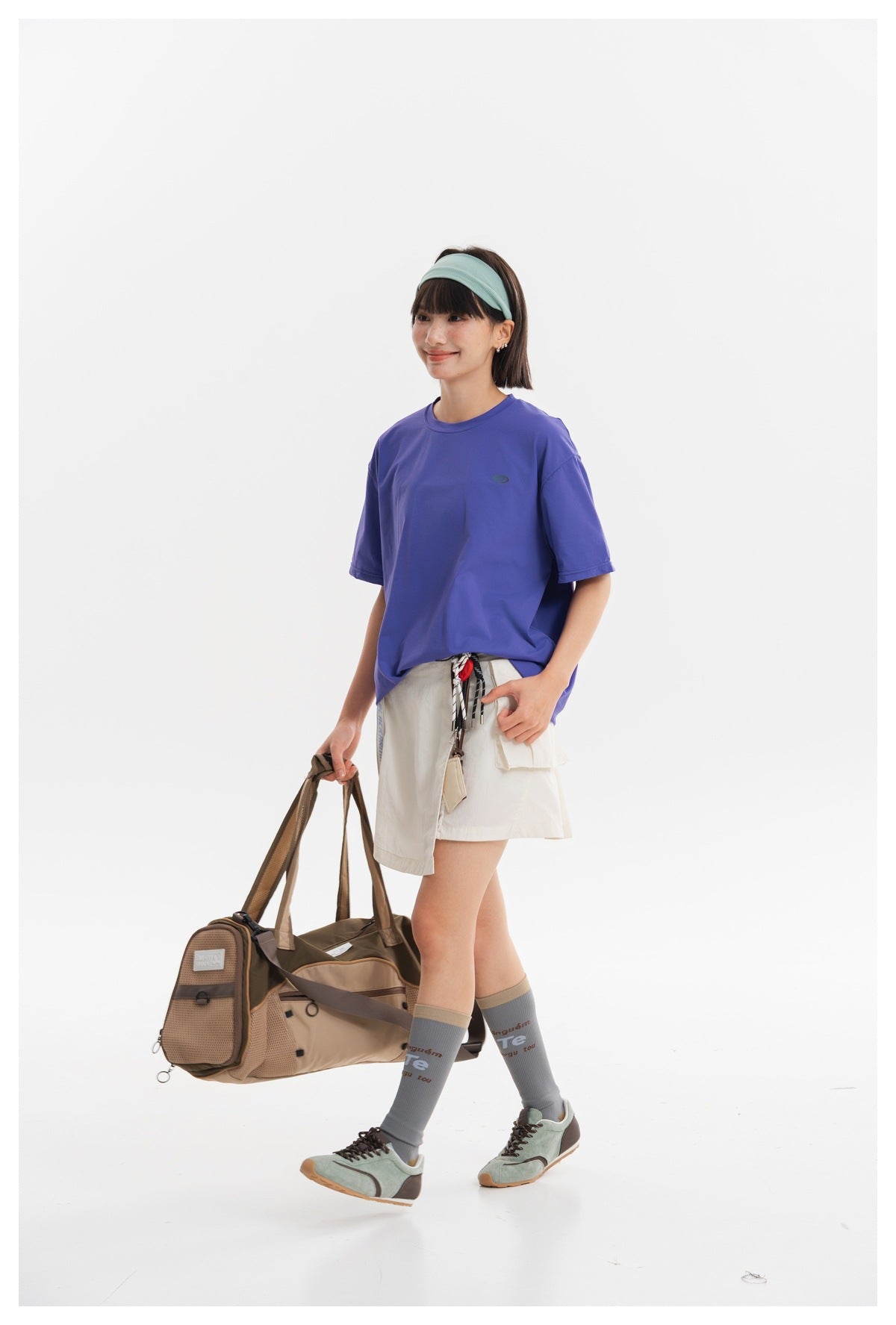 Model wearing an off-white utility skirt and a loose purple T-shirt, carrying a brown duffel bag.