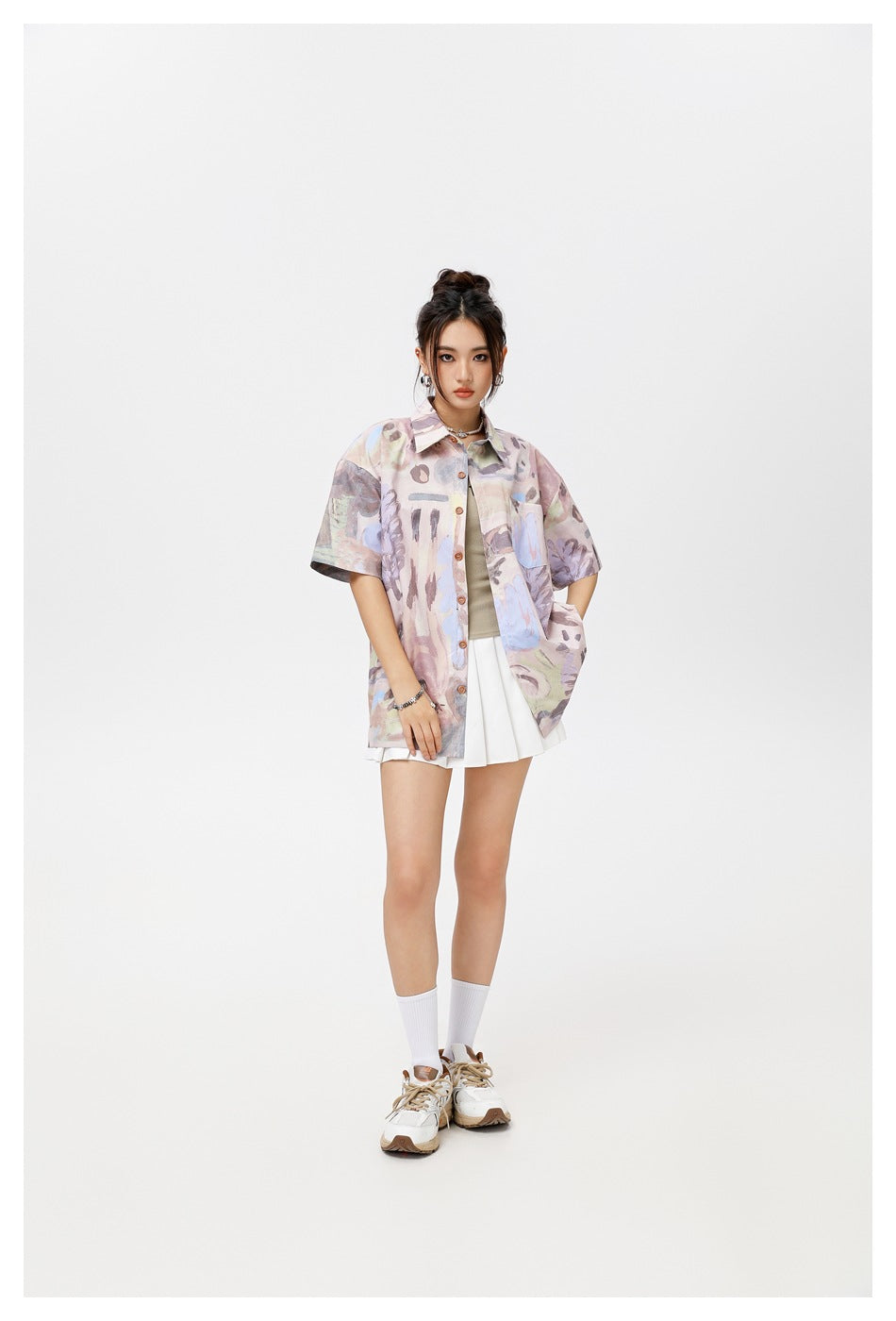 Model wearing pink-purple abstract print short-sleeve shirt open over a tan top and white pleated skirt