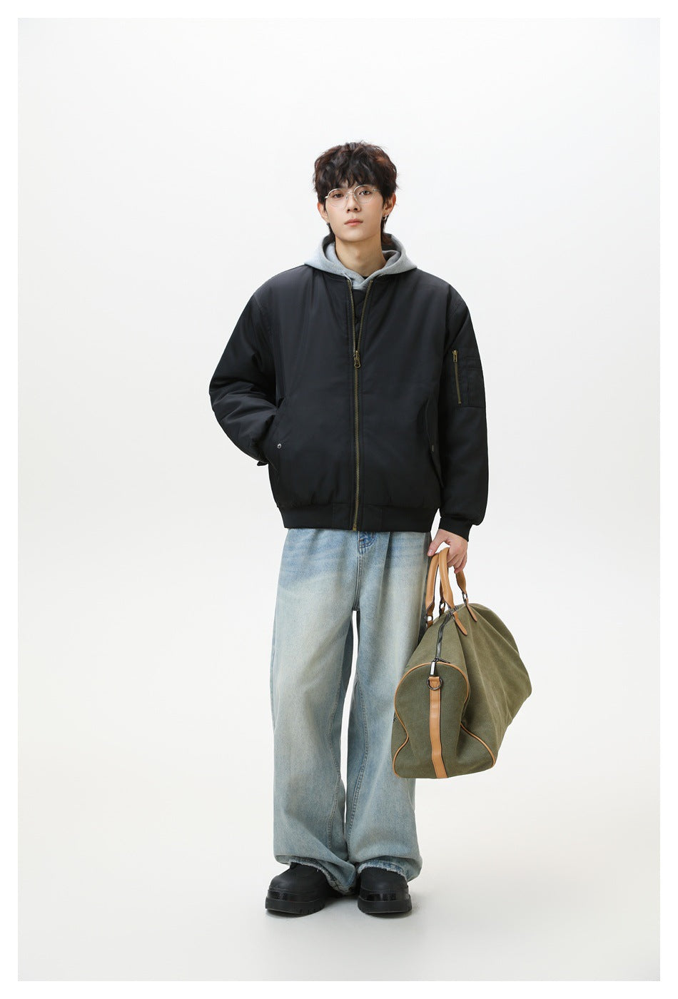 Full-length model wearing a black bomber jacket over a gray hoodie, with a large olive bag.