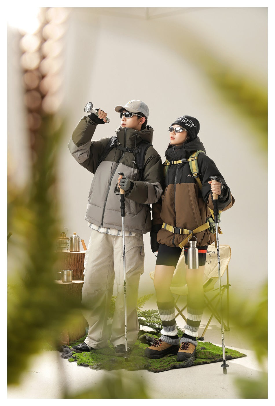 Two models wear olive-gray and black-brown color-block puffer jackets with backpacks; blurred foliage frames the camping