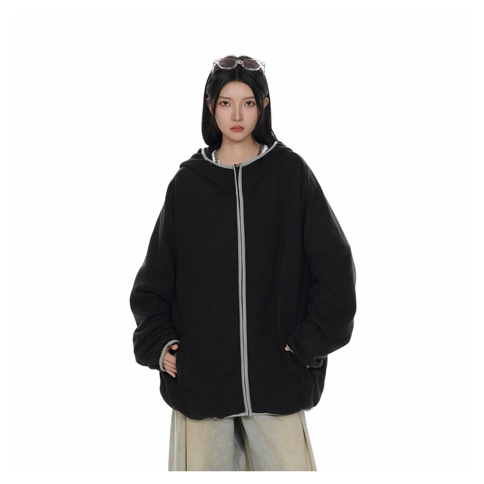 Model wearing an oversized black hooded zip jacket closed, with gray piping, hands in pockets, and light wide-leg pants