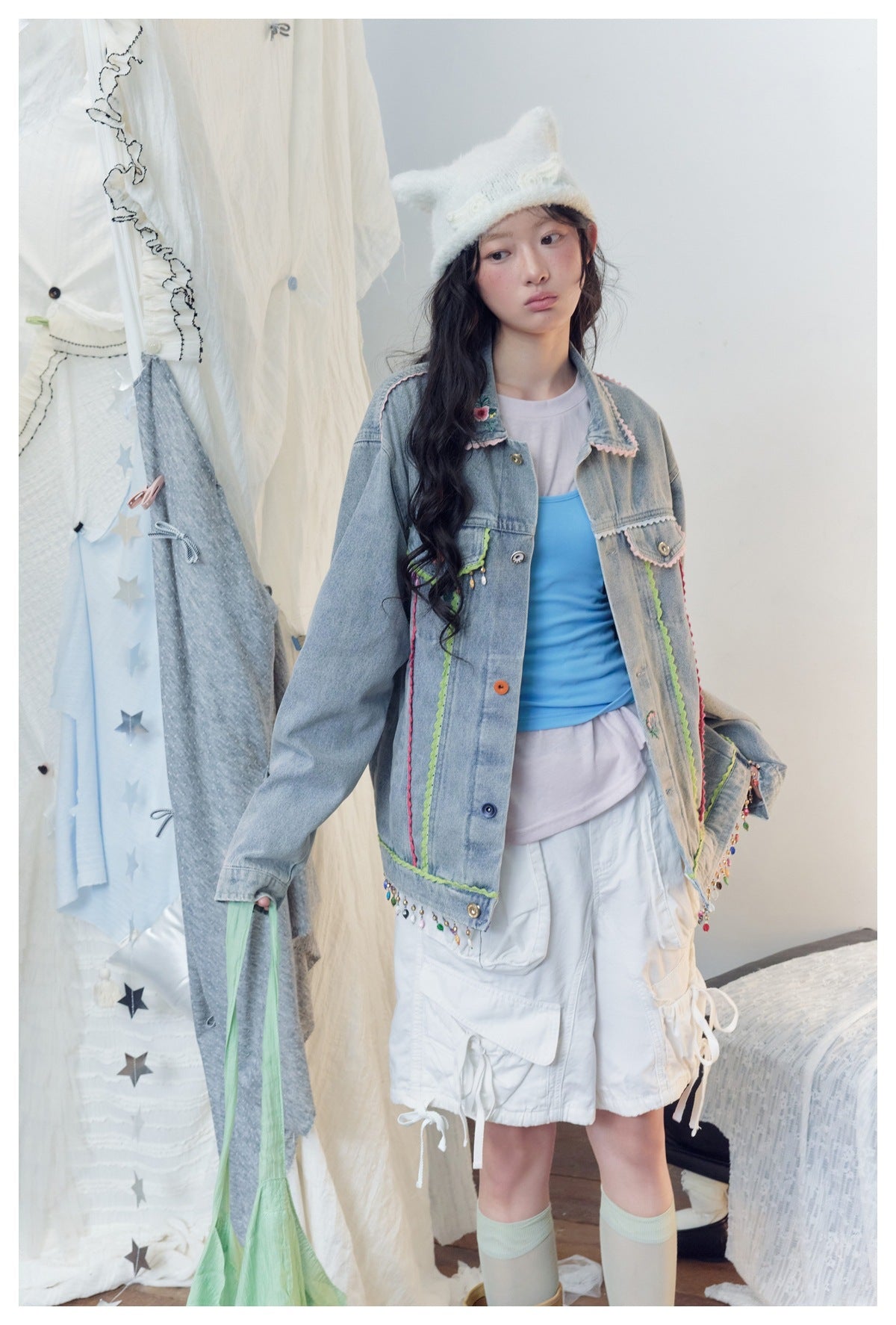 Model wearing a light-blue denim jacket with colorful trim and dangling beads, paired with white cargo shorts.
