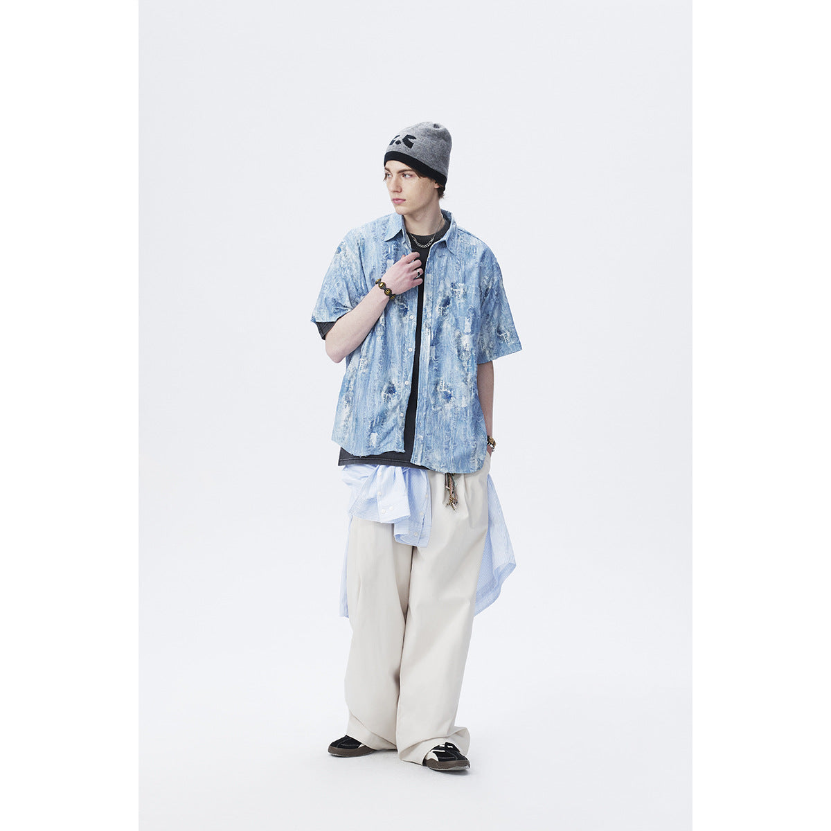 Full-length model wearing a blue distressed-pattern short-sleeve button-up over a black tee with loose cream trousers.