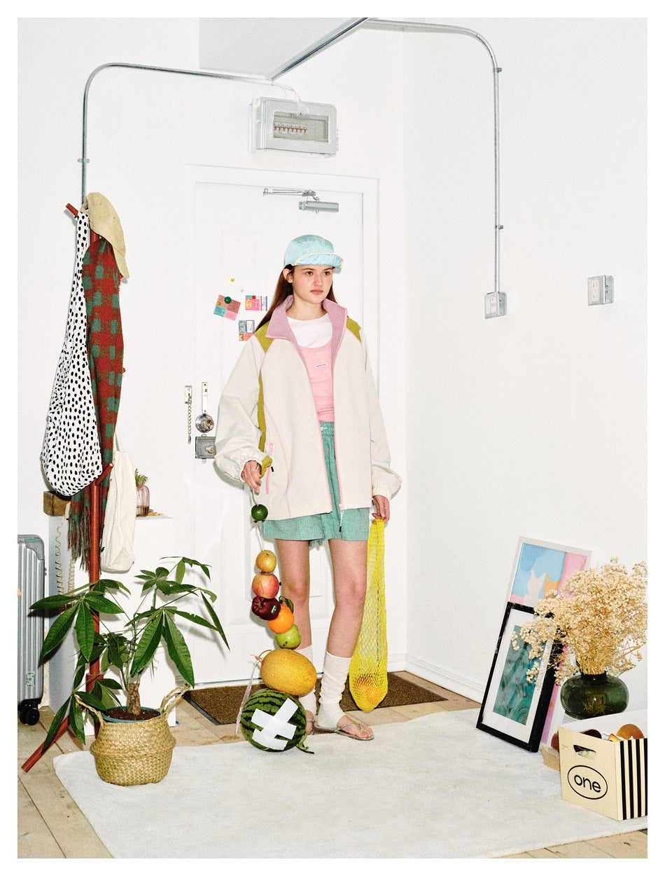 Full-length model wearing an ivory color-block jacket with pink trim, turquoise shorts and a pale blue cap.