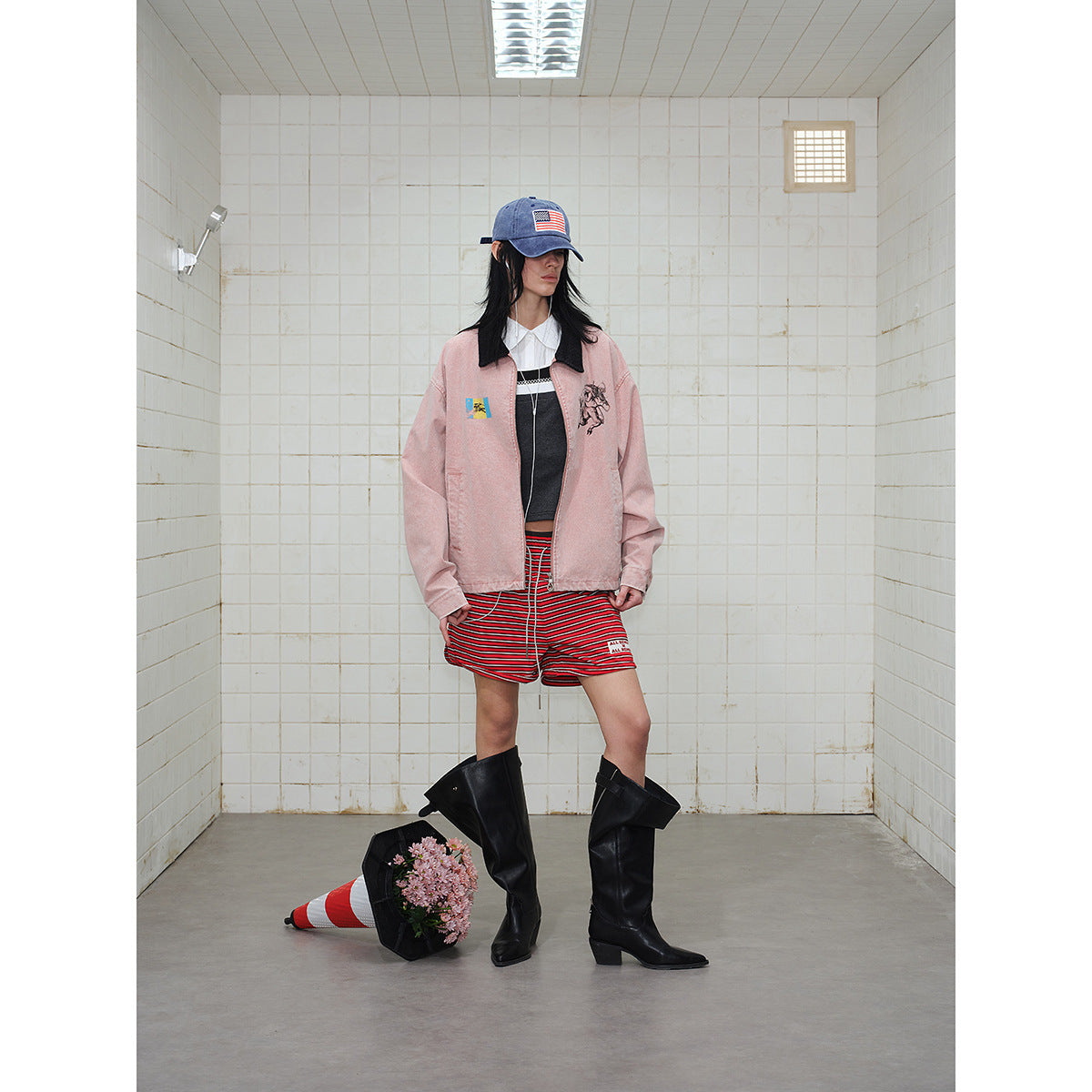 Full-length model wearing a dusty pink graphic jacket, red striped shorts, blue cap, and tall black boots.