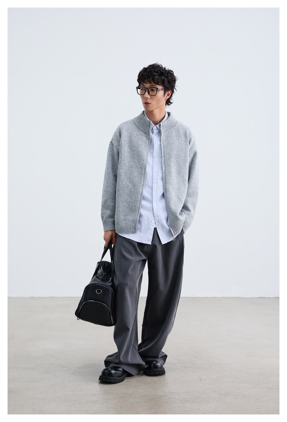 Full-length model wearing an open light gray zip cardigan over a striped shirt and dark trousers.