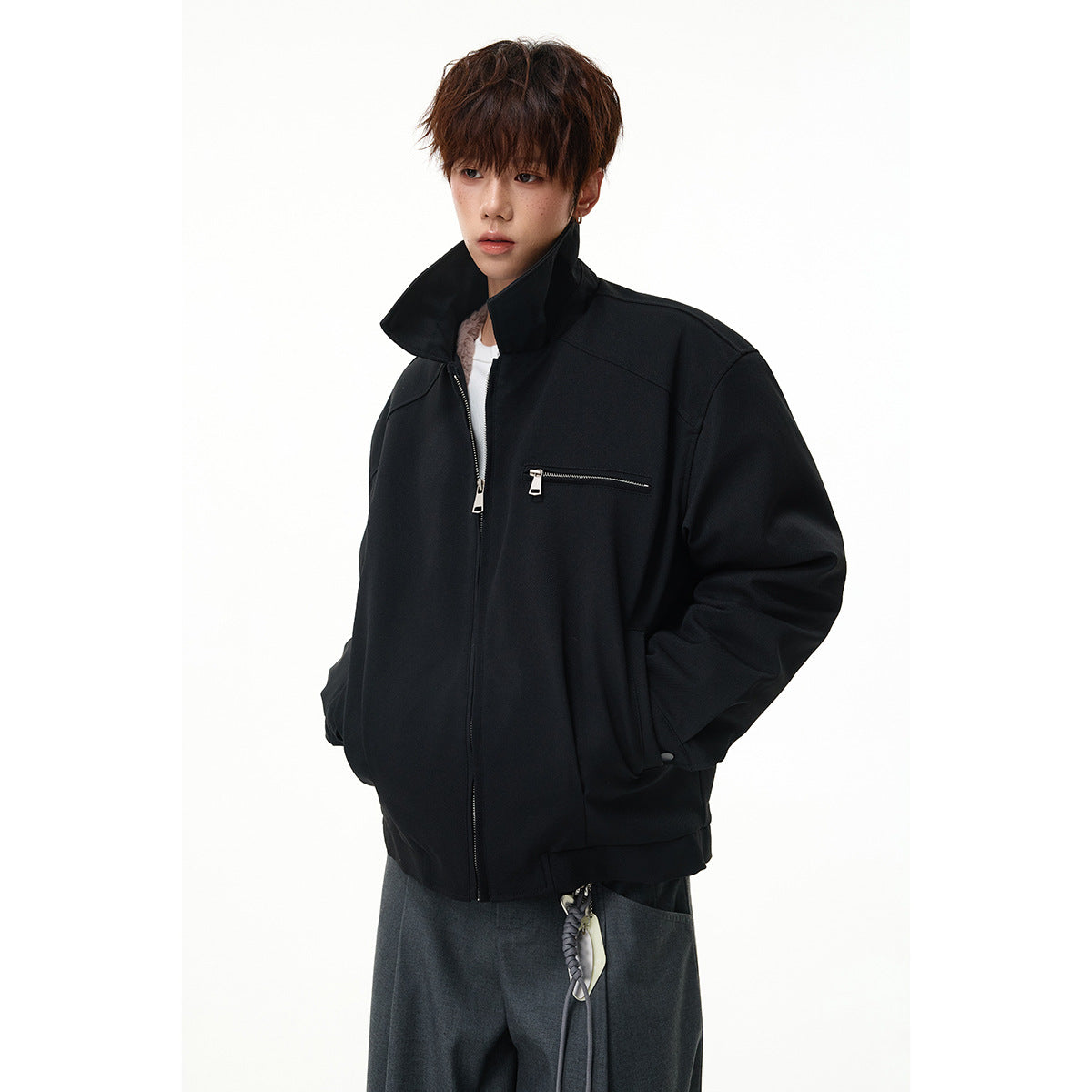 Model wearing a black zip jacket with a raised collar and chest zip pocket, over gray trousers.