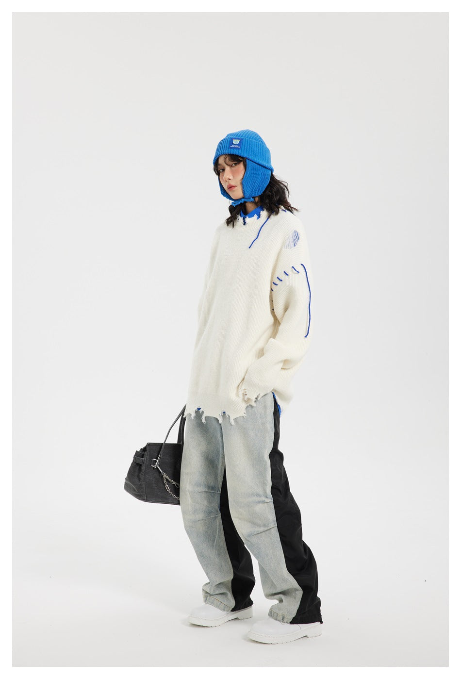Model wearing an oversized distressed ivory sweater with blue stitch accents, a bright blue earflap beanie, and loose jeans.