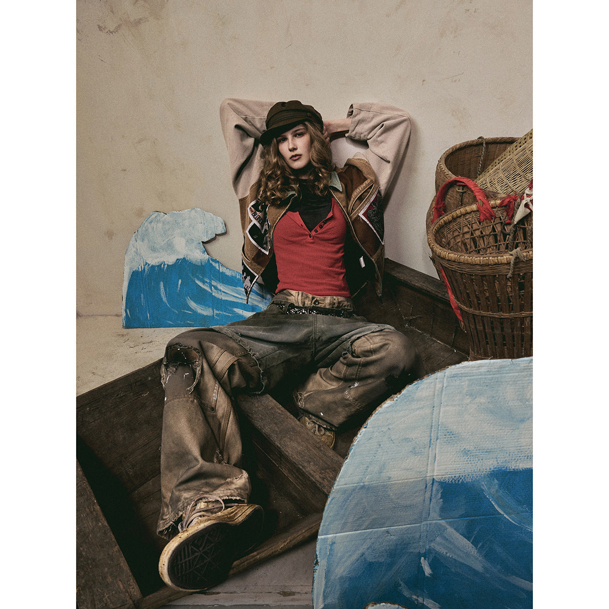 Model seated in a brown patchwork jacket with pale sleeves, red top, dark cap, and loose gray-brown pants.
