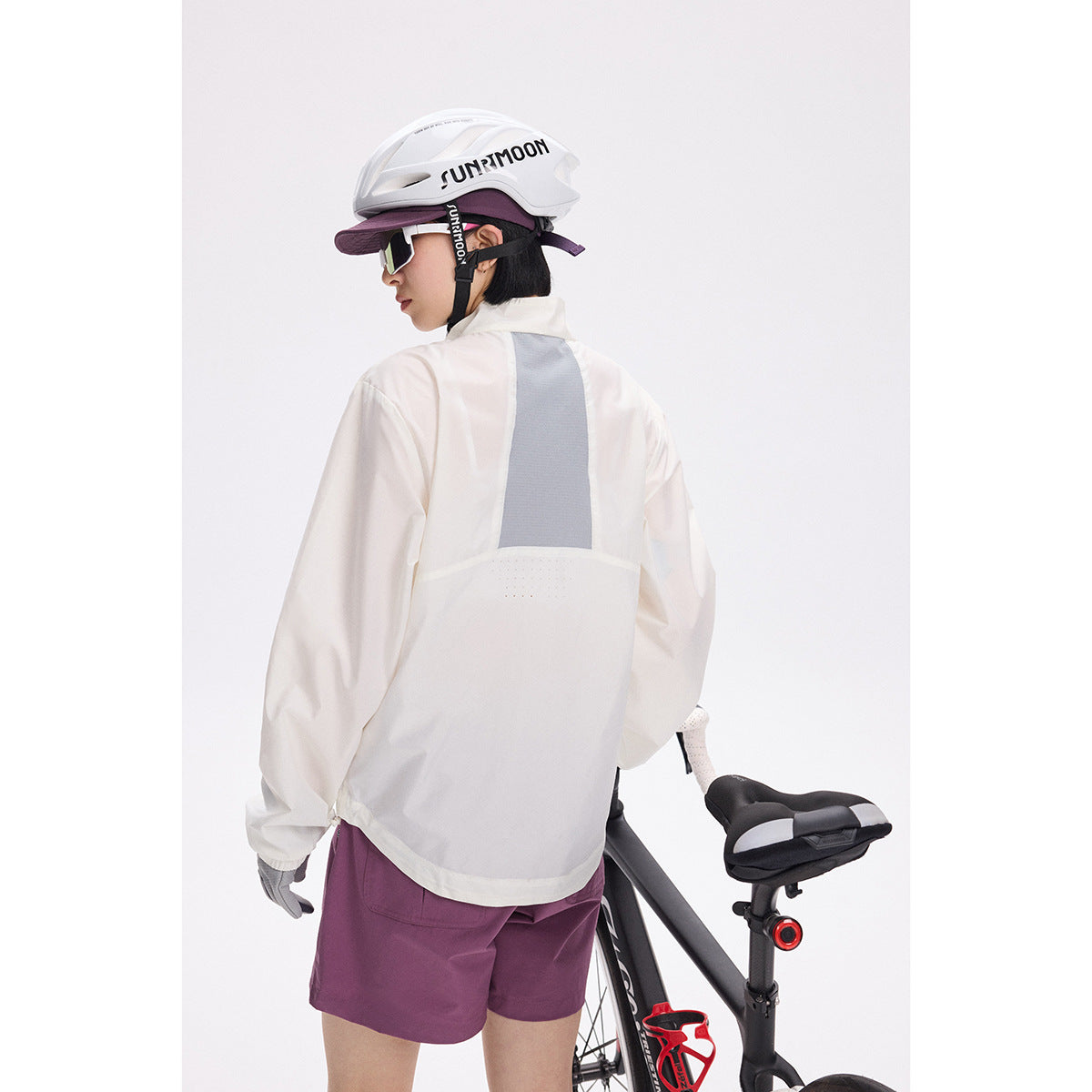 Rear view of a cyclist wearing an ivory jacket with a gray back panel and plum shorts beside a road bike.