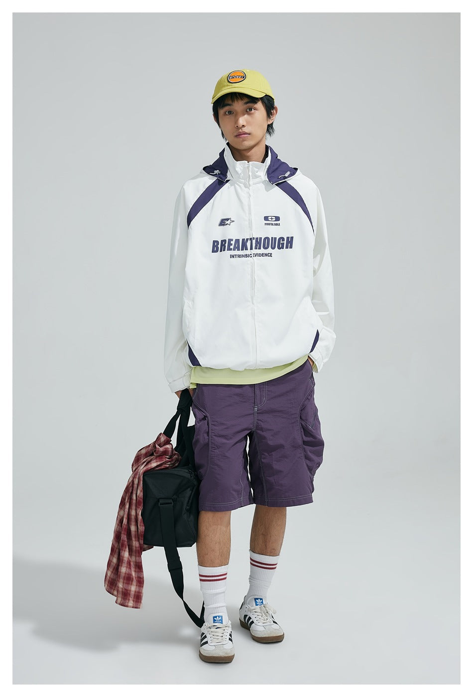 Model wearing white hooded zip jacket with purple panels, purple shorts and a yellow cap, standing full length