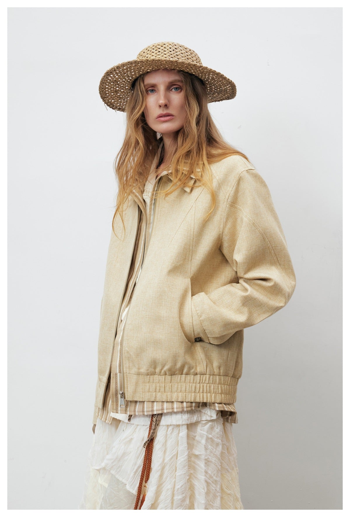 Model wearing khaki zip-front jacket over layered white clothing, side view with hand in pocket and straw hat