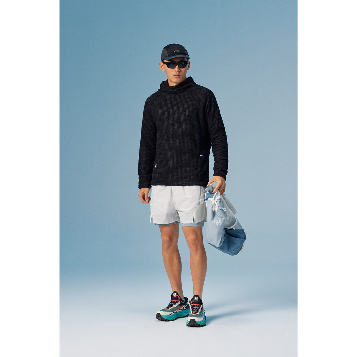 Model wearing a black textured high-neck pullover and white shorts, carrying a blue bag against a blue backdrop.