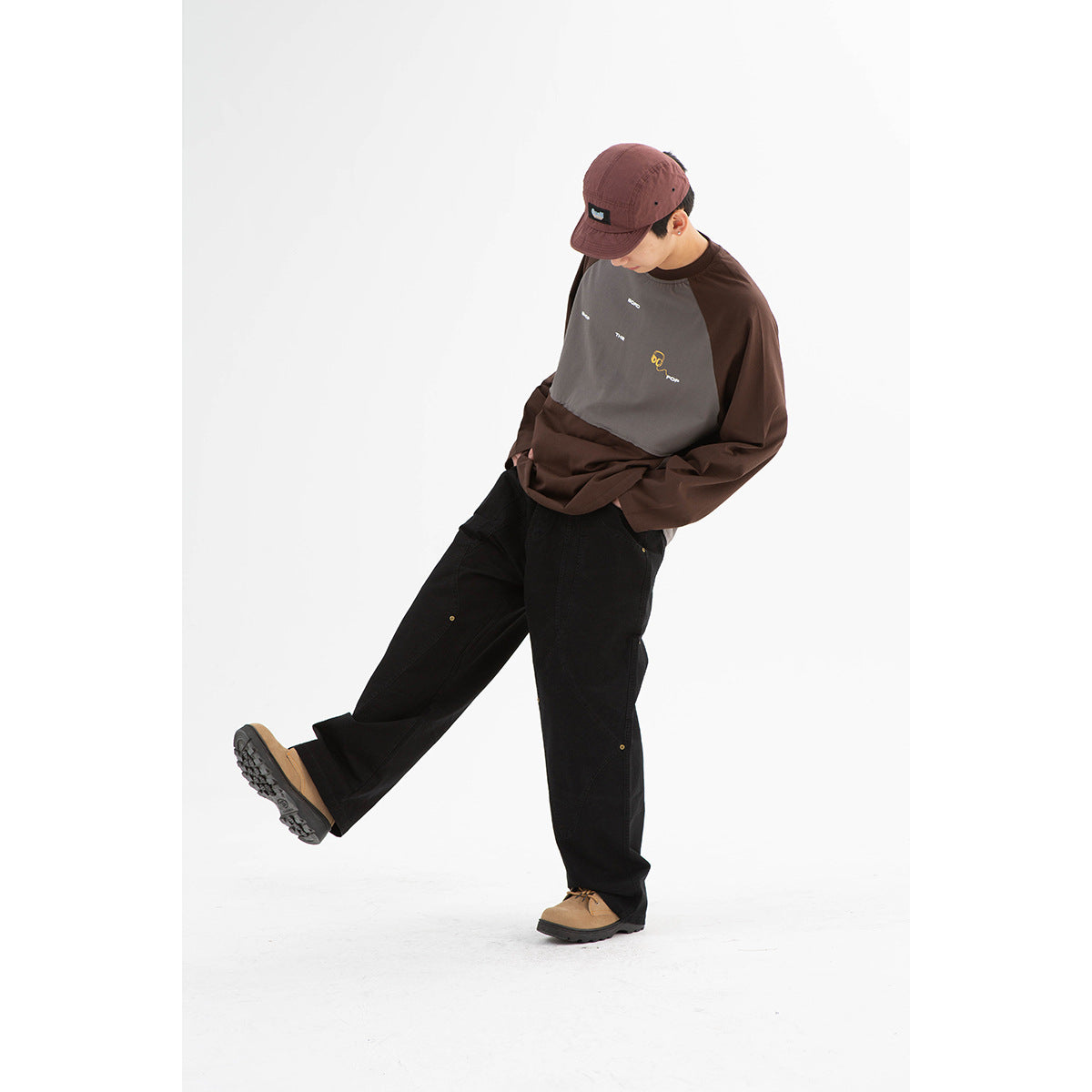 Model wearing a brown and gray color-block raglan sweatshirt with chest graphics, black trousers, and a burgundy cap.