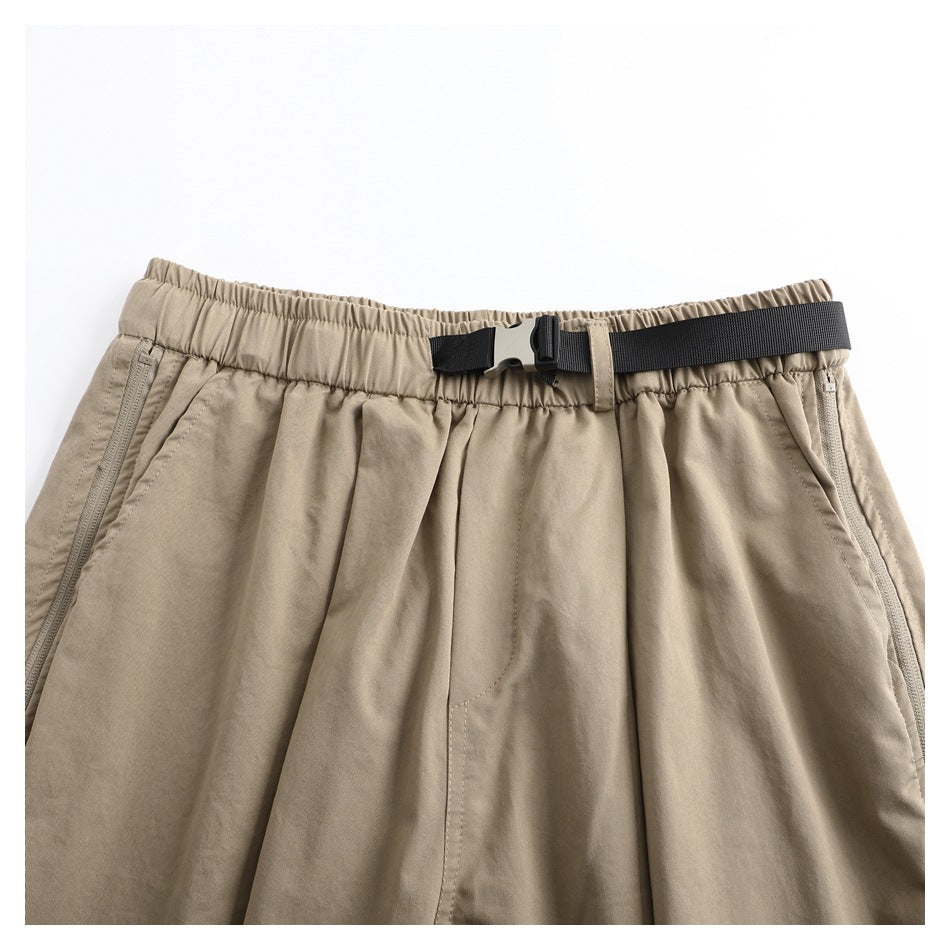 Close view of the front waist of taupe trousers with an elastic waistband, black webbing belt and side zip pockets.