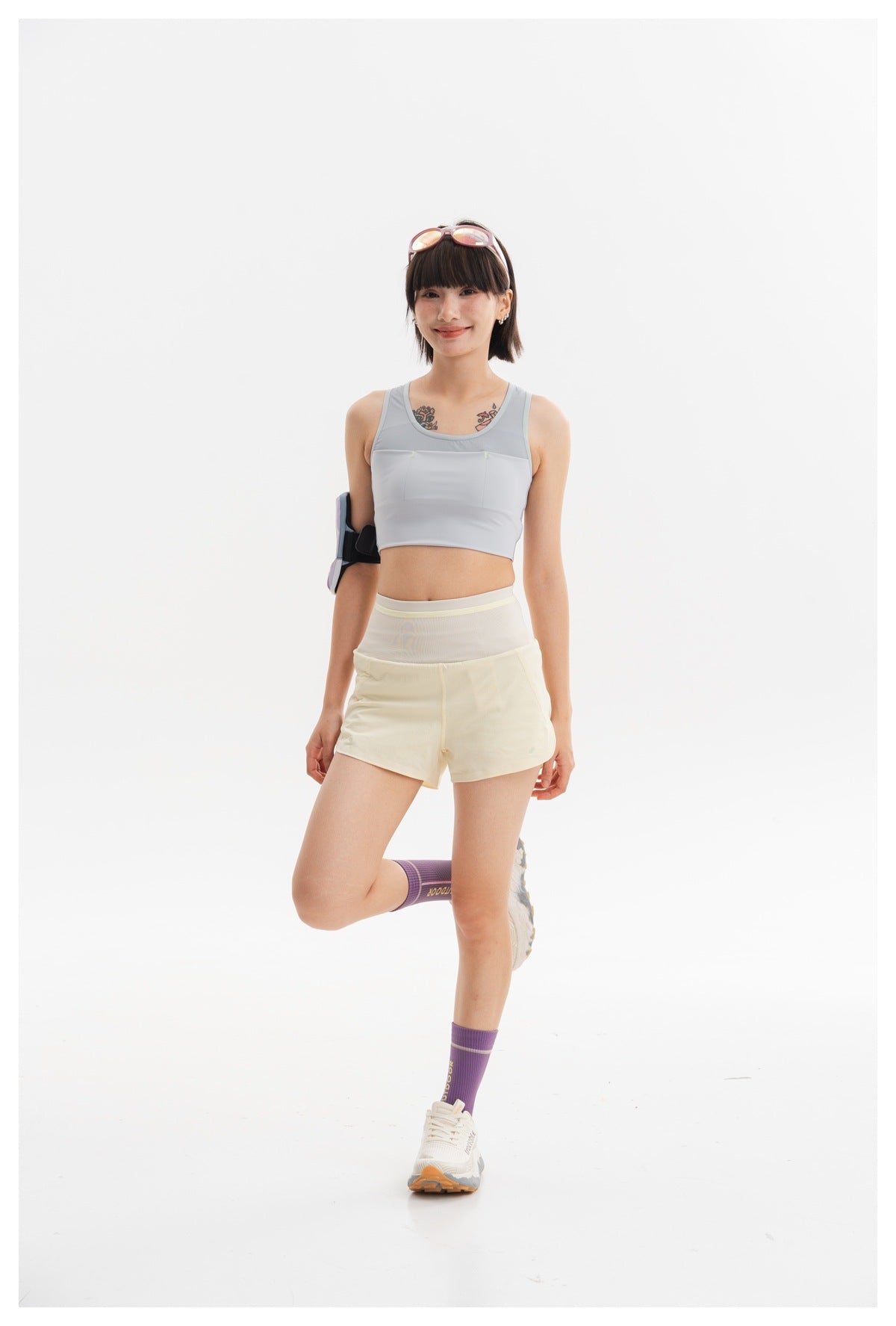 Model in a light gray cropped athletic tank and pale yellow shorts, posing with one leg bent and a padded arm accessory.