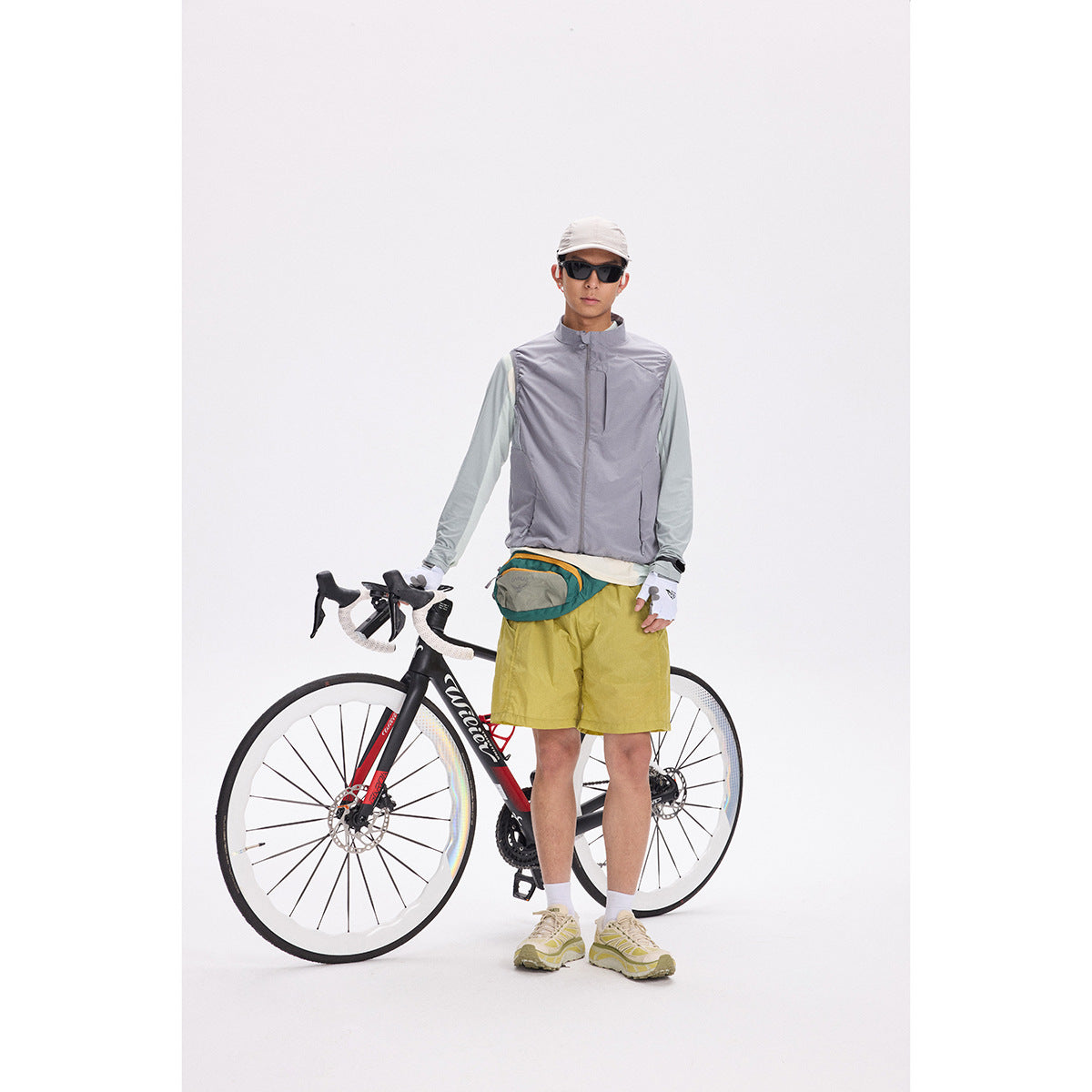 Model wearing a gray zip vest over a pale blue long-sleeve top, standing beside a road bicycle.