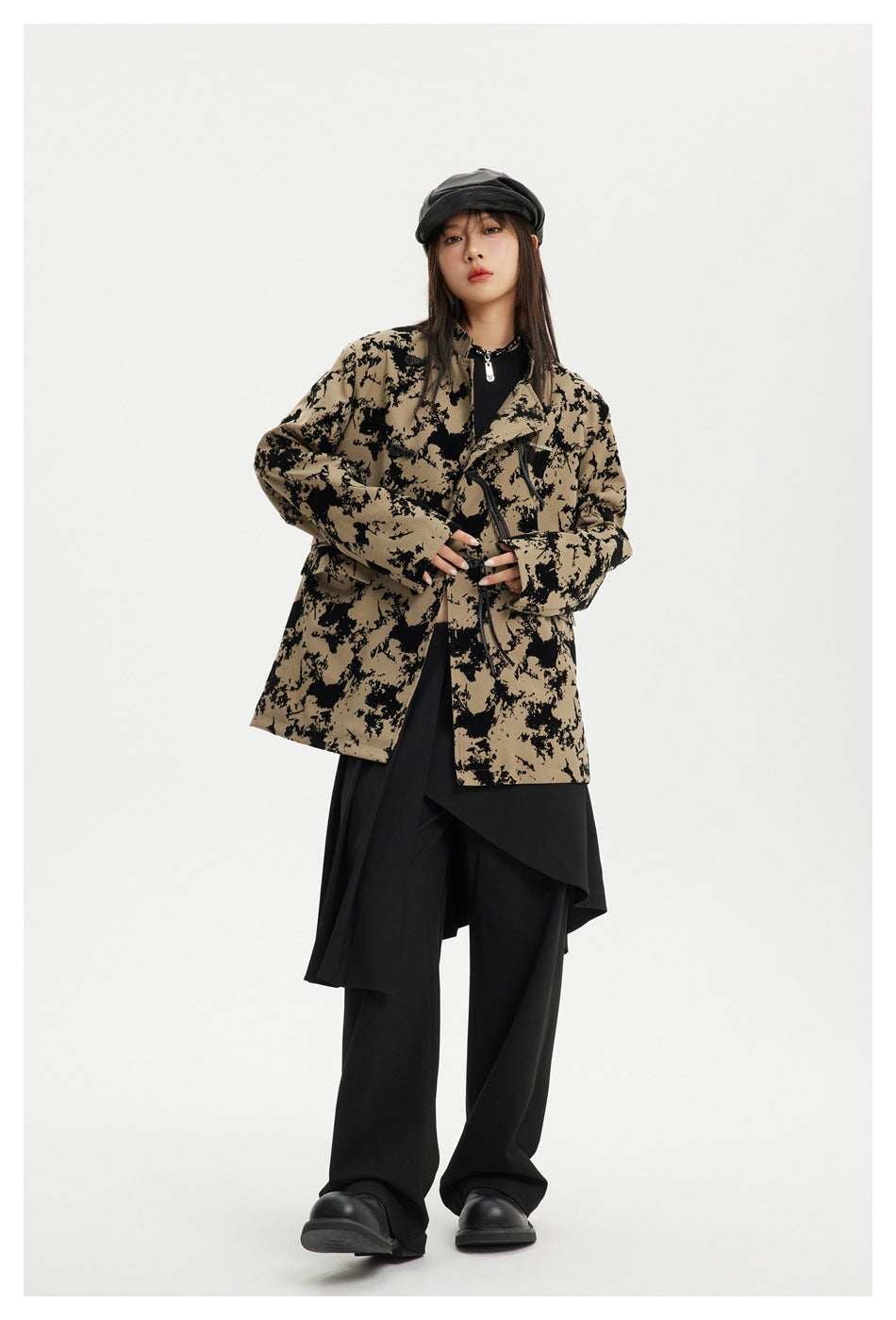 Full-length model wearing a tan-and-black abstract-patterned toggle jacket with wide black trousers and a cap.