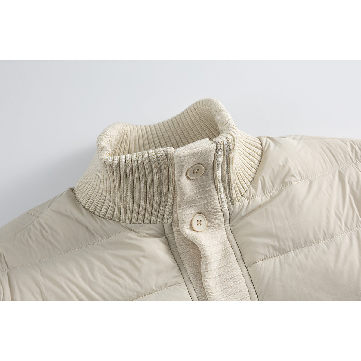 Close-up of a cream jacket’s ribbed stand collar and buttoned knit placket beside its quilted body.