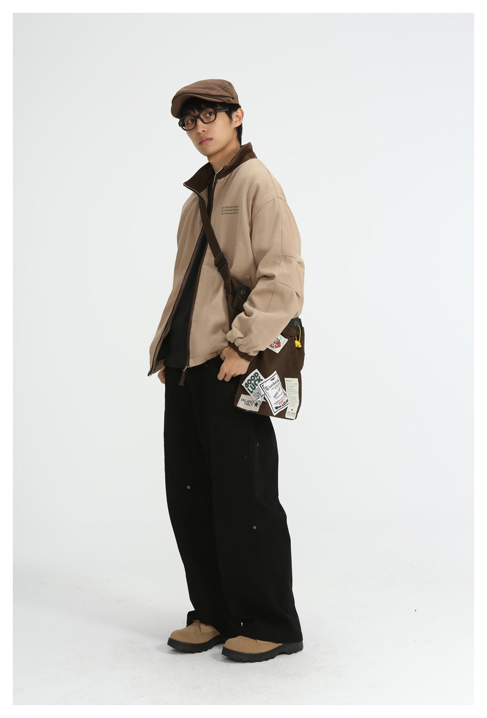 Model in a khaki stand-collar jacket with brown cuffs, seen at an angle with black trousers, cap and a patched crossbody bag