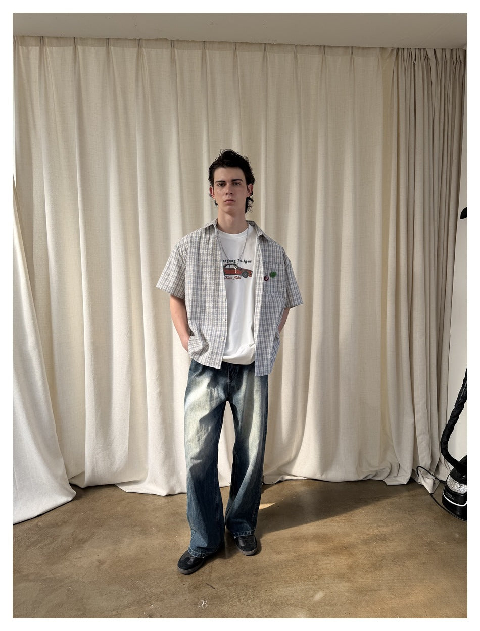 Full-length front view of a man wearing an open blue-gray plaid short-sleeve shirt, white graphic tee, and loose dark jeans.