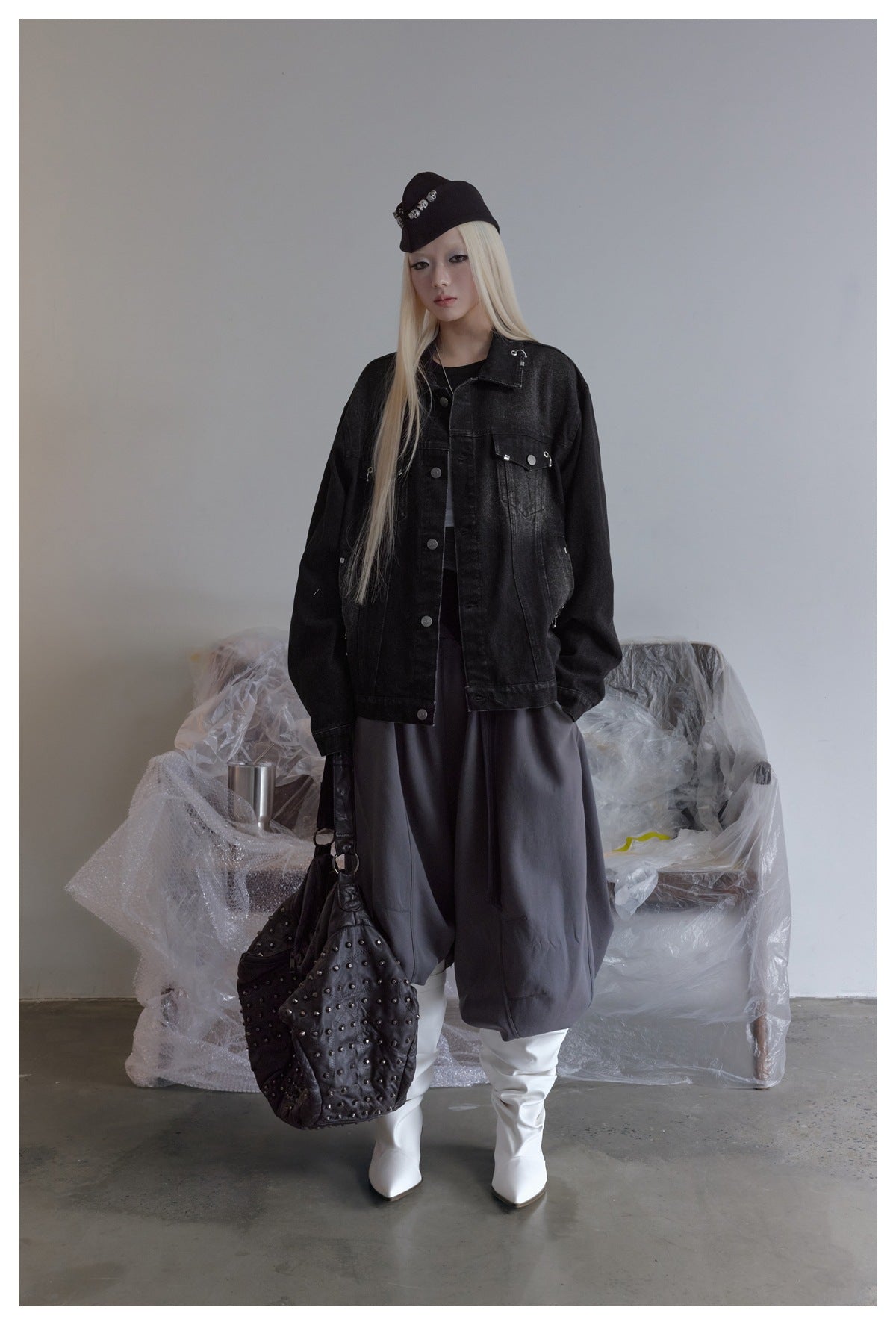 Model wearing open black washed denim jacket with metal hardware over dark top, gray pants and white boots