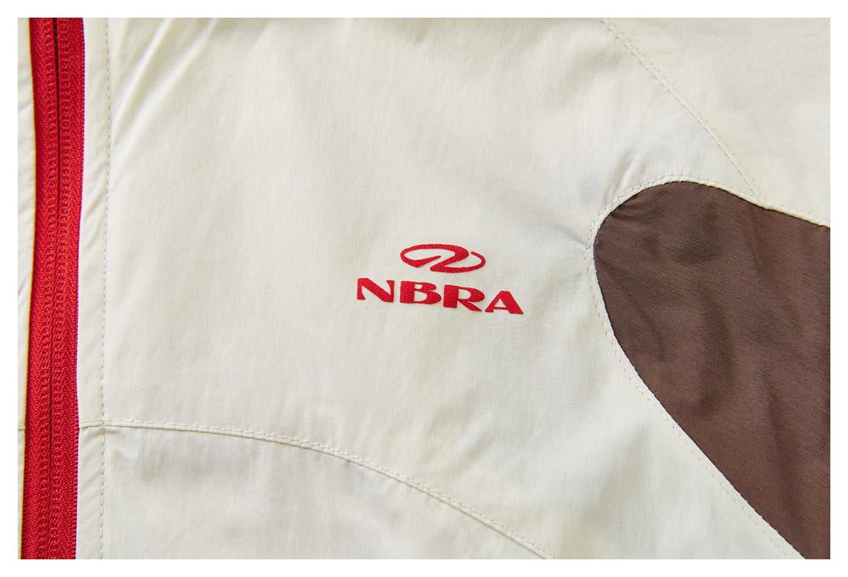 Close-up of a cream garment with a red embroidered NBRA logo, red zipper, and brown contrast panel.