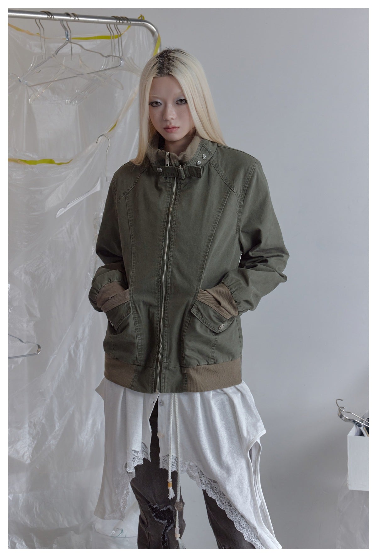 Front view of a model wearing an olive-gray zip jacket with a buckled stand collar, brown ribbed trim, and flap pockets.