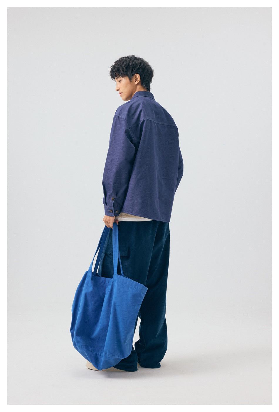 Back view of a model in a blue long-sleeve shirt with a back yoke seam, holding a blue tote bag