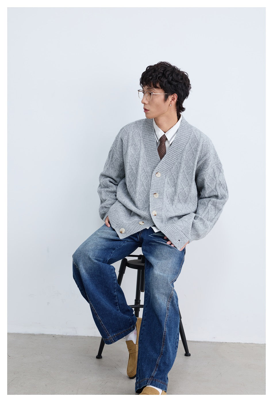 Man seated on a stool wearing a light gray cable-knit cardigan over a white shirt and tie with wide blue jeans