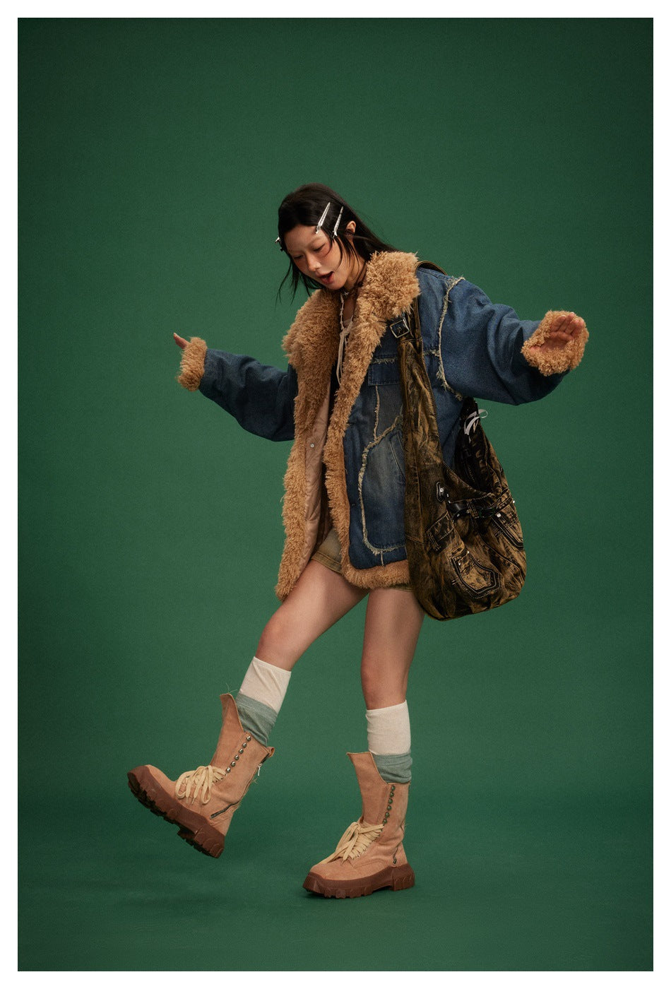 Model wearing the navy denim jacket with tan fleece-like trim over a short skirt, with boots and a large bag, on a green