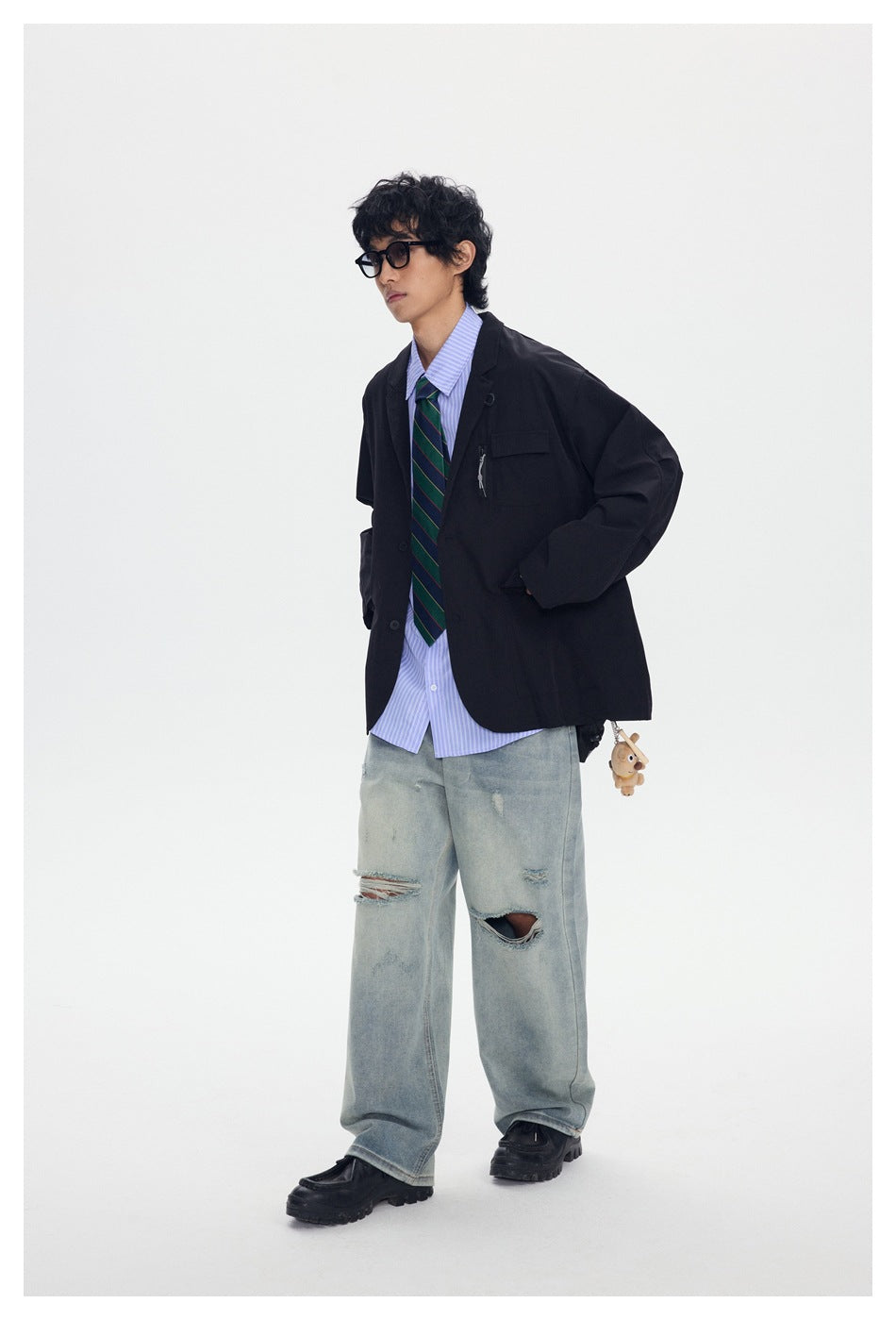 Full-length view of a model in a black oversized blazer, striped shirt and tie, loose distressed jeans, and chunky shoes.