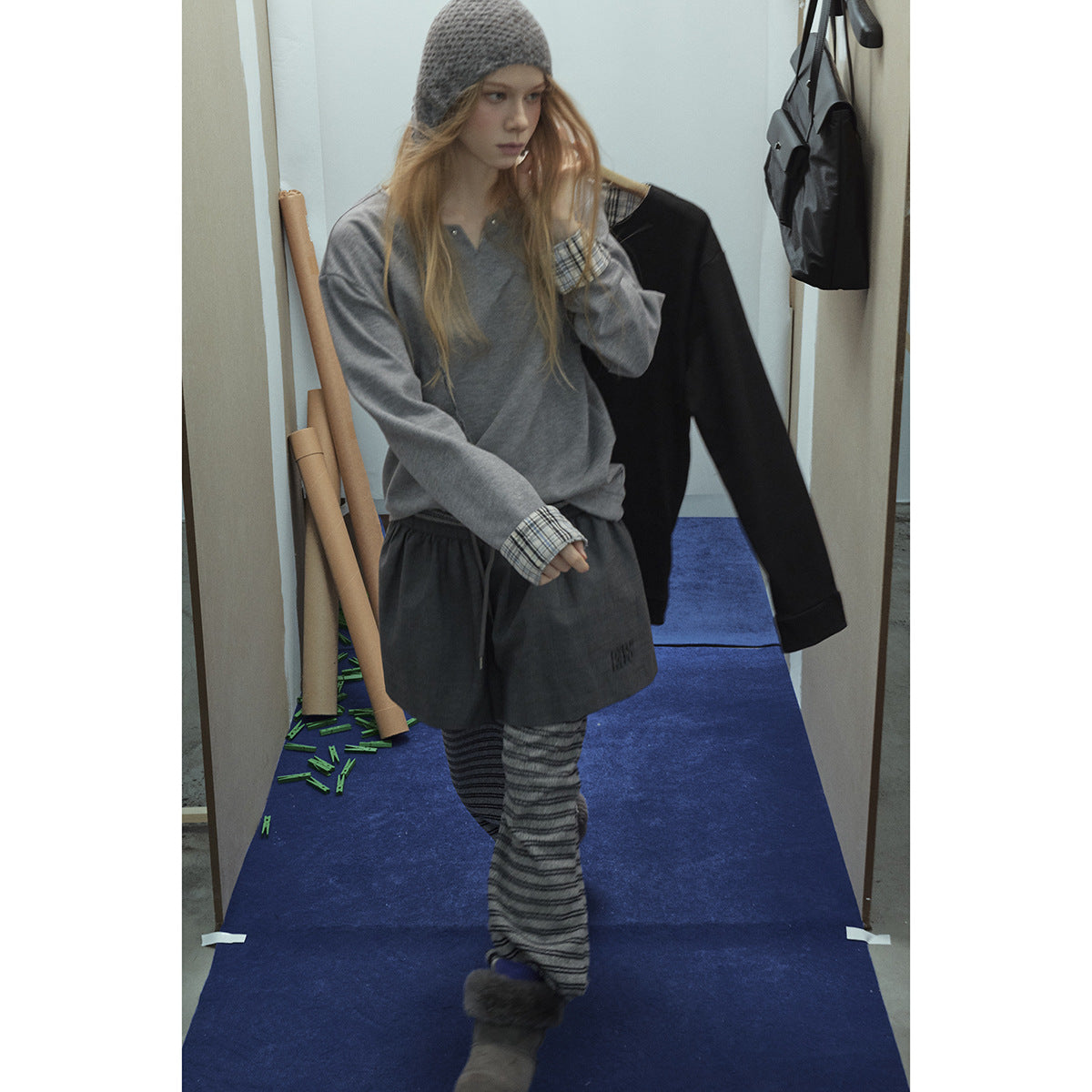 Model in a gray pullover with plaid cuffs, dark shorts over striped trousers, and a gray knit hat.