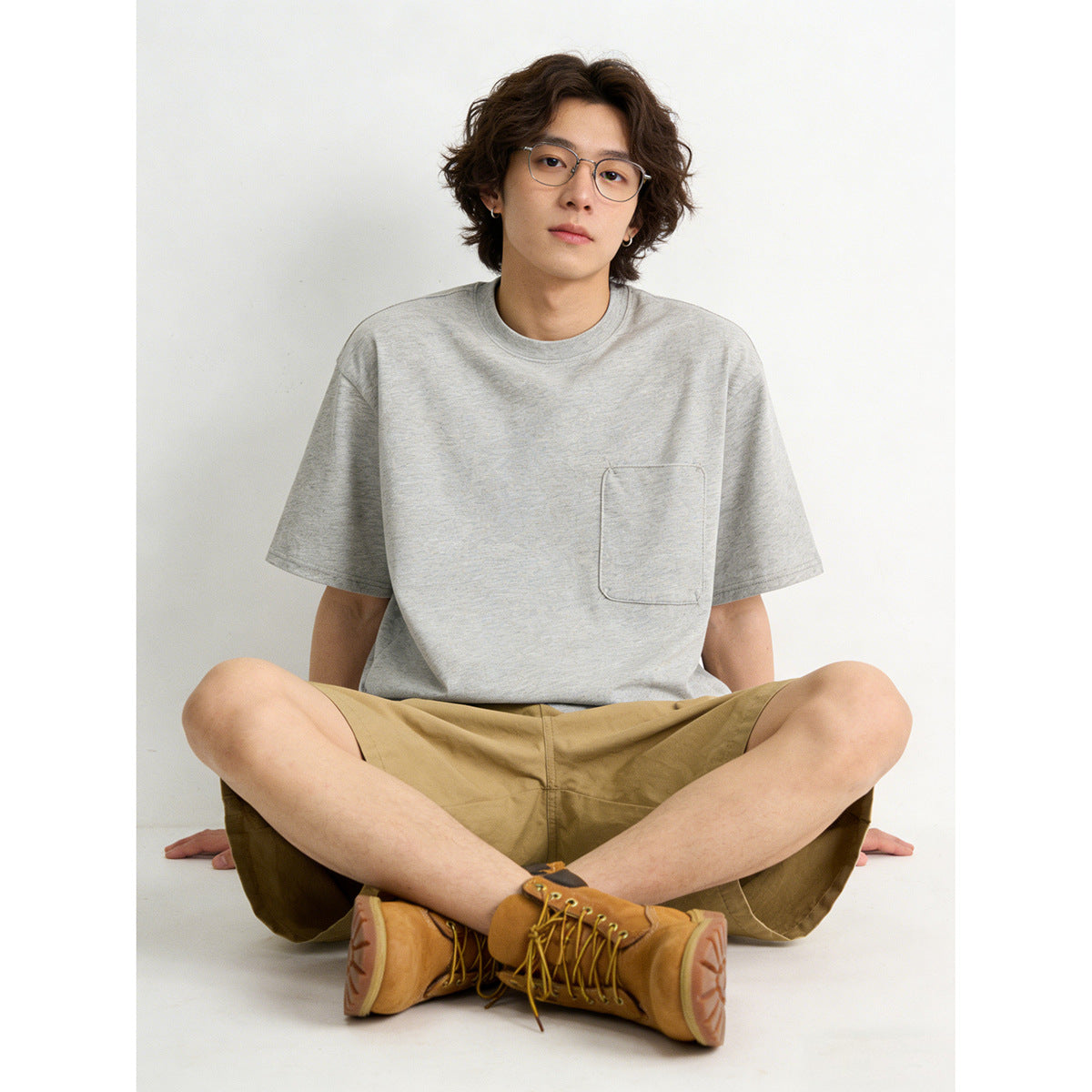 Seated model wearing a light gray crew-neck T-shirt with a chest pocket and tan shorts.