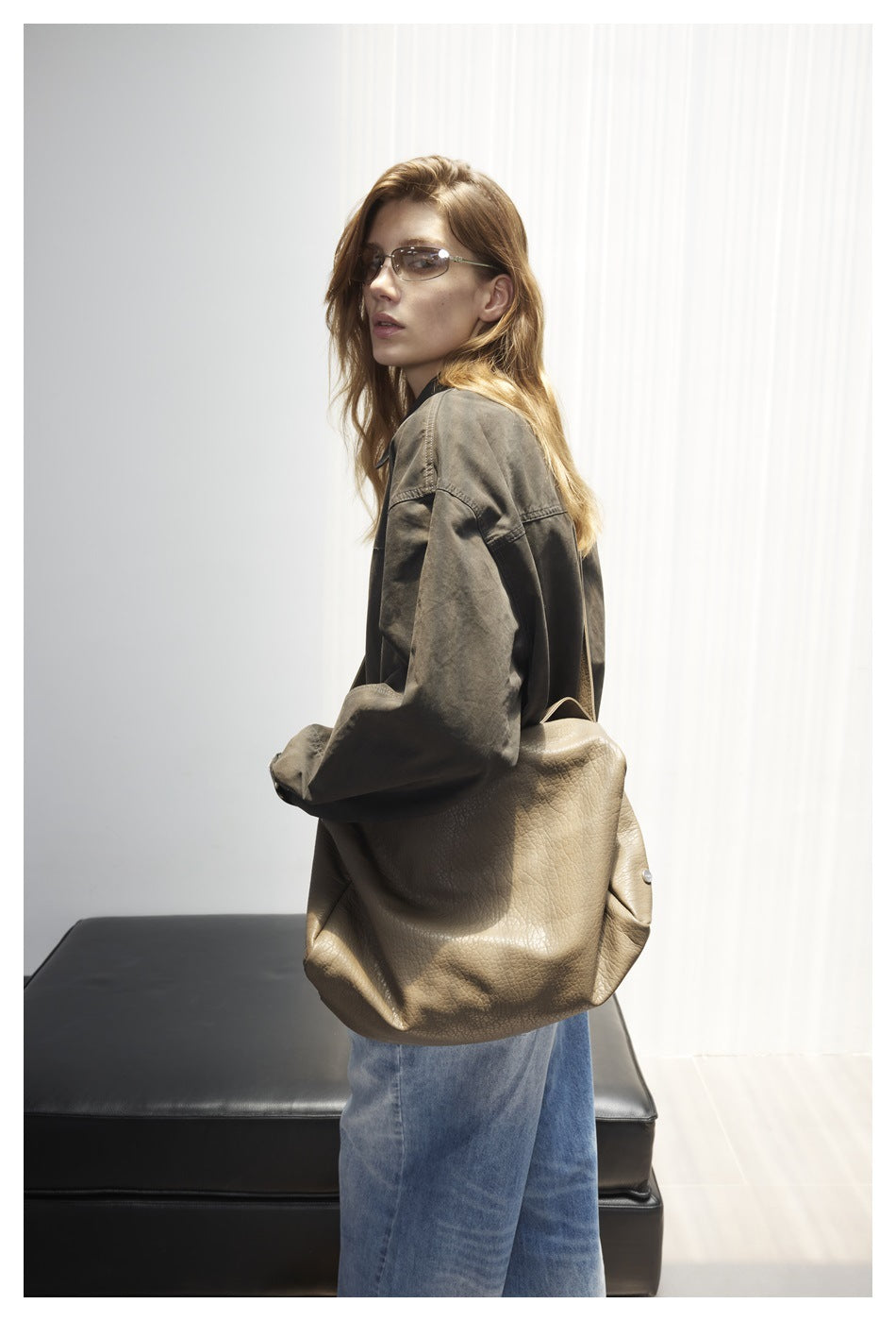 Model in a dark olive oversized jacket, shown from the side with a large taupe shoulder bag and wide-leg blue jeans.