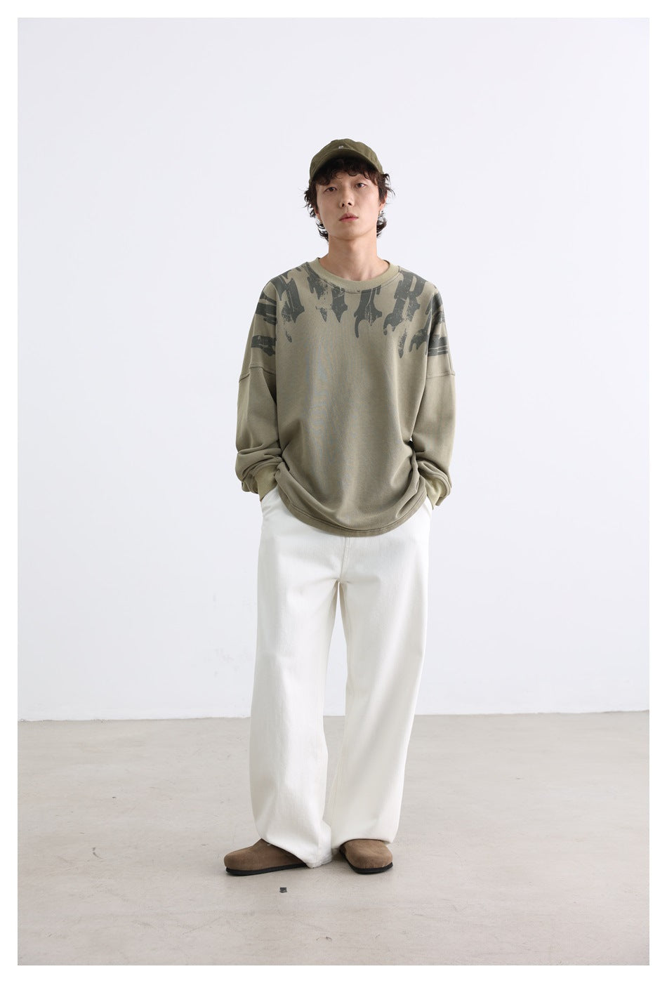 Model wearing an olive oversized crewneck sweatshirt with dark shoulder graphics and white wide-leg pants.