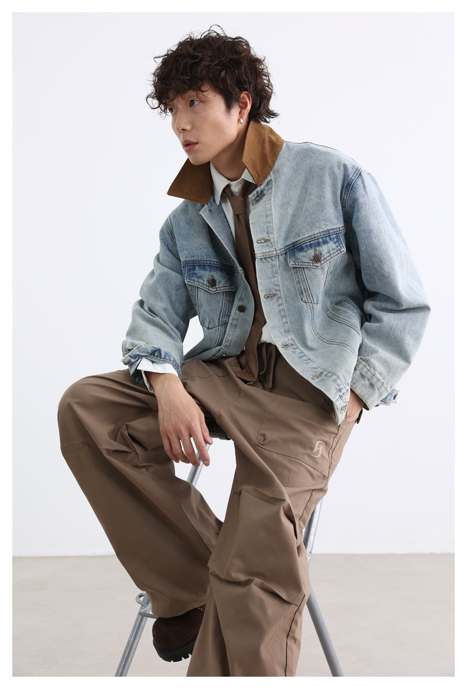 Seated model wearing a light-wash denim jacket with a brown collar and loose brown trousers.