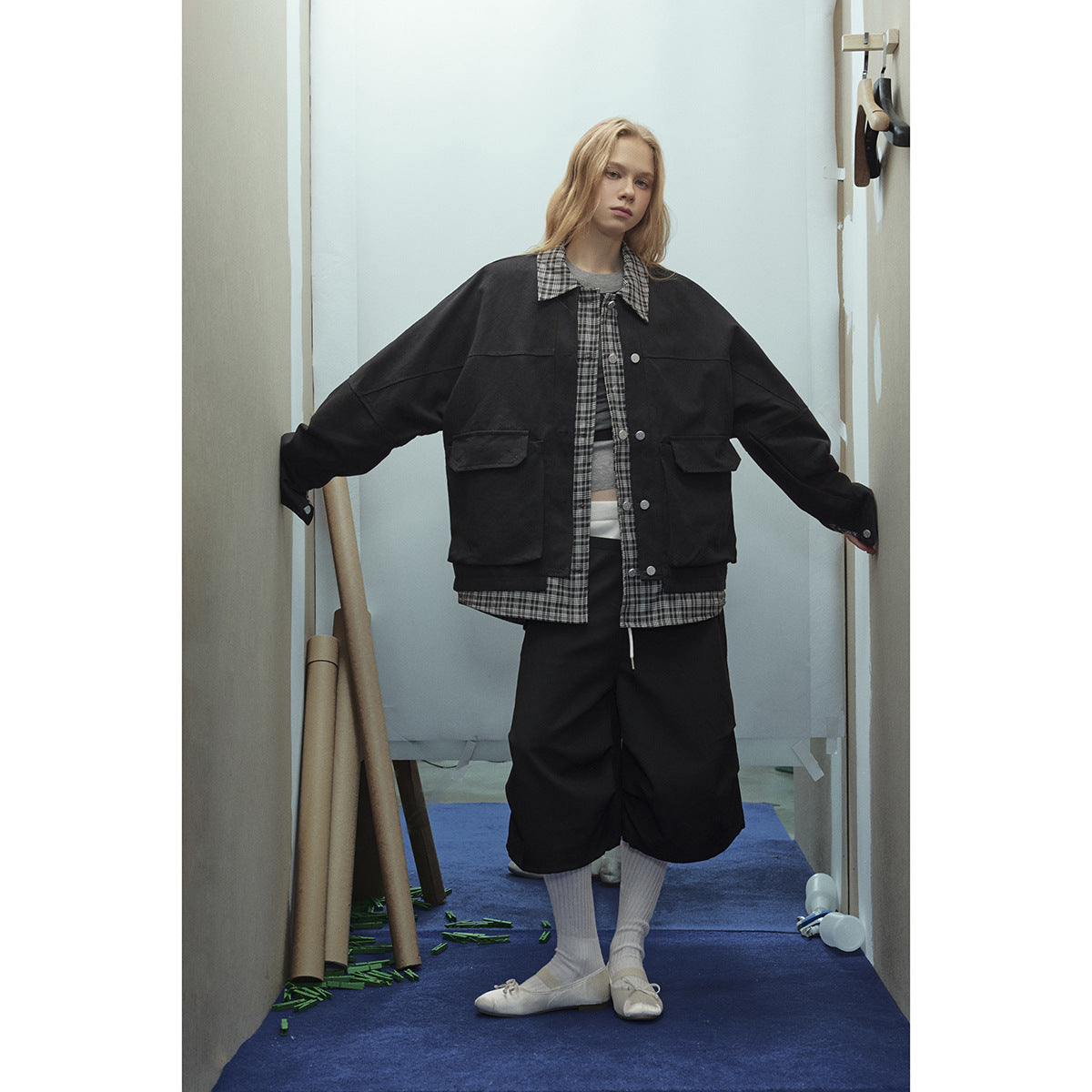 Front view of a model in an oversized black jacket with plaid trim and large flap pockets, paired with black shorts.