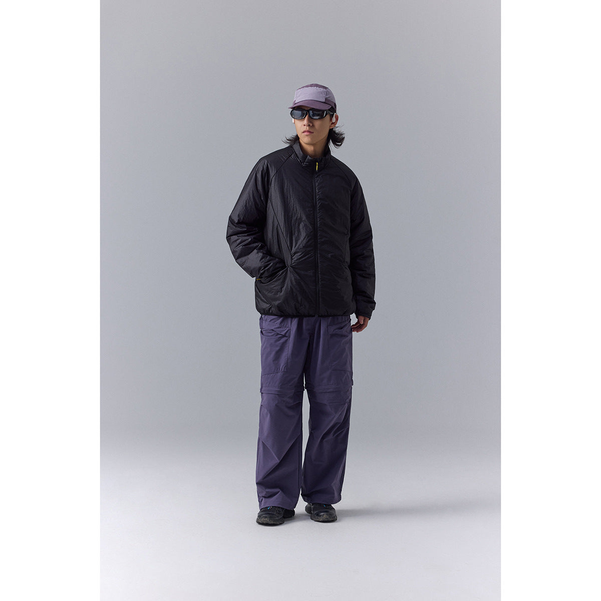 Front view of a model wearing a black zip jacket, loose purple pants, a cap, and sunglasses.