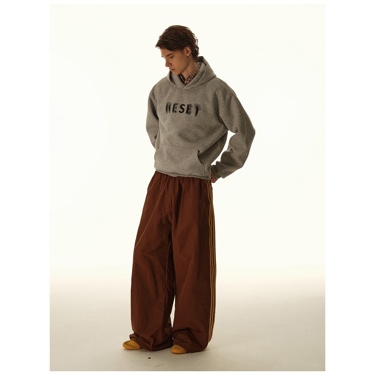 Model wearing a gray hooded sweatshirt with dark chest lettering and wide brown striped pants.