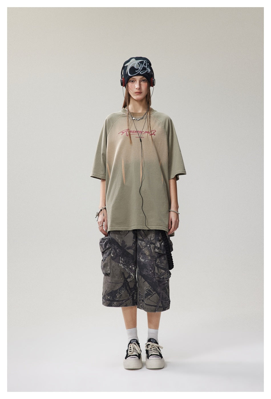 Full-length front view of a model wearing an oversized sage-green graphic T-shirt and dark camouflage cargo shorts.