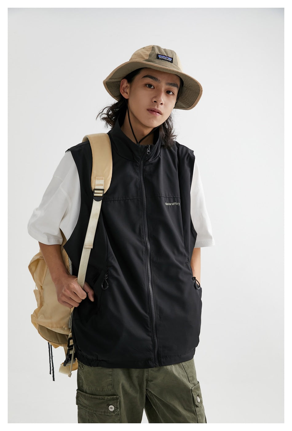 Front view of a model wearing a black sleeveless zip vest over a white T-shirt, with olive cargo pants and a tan hat.