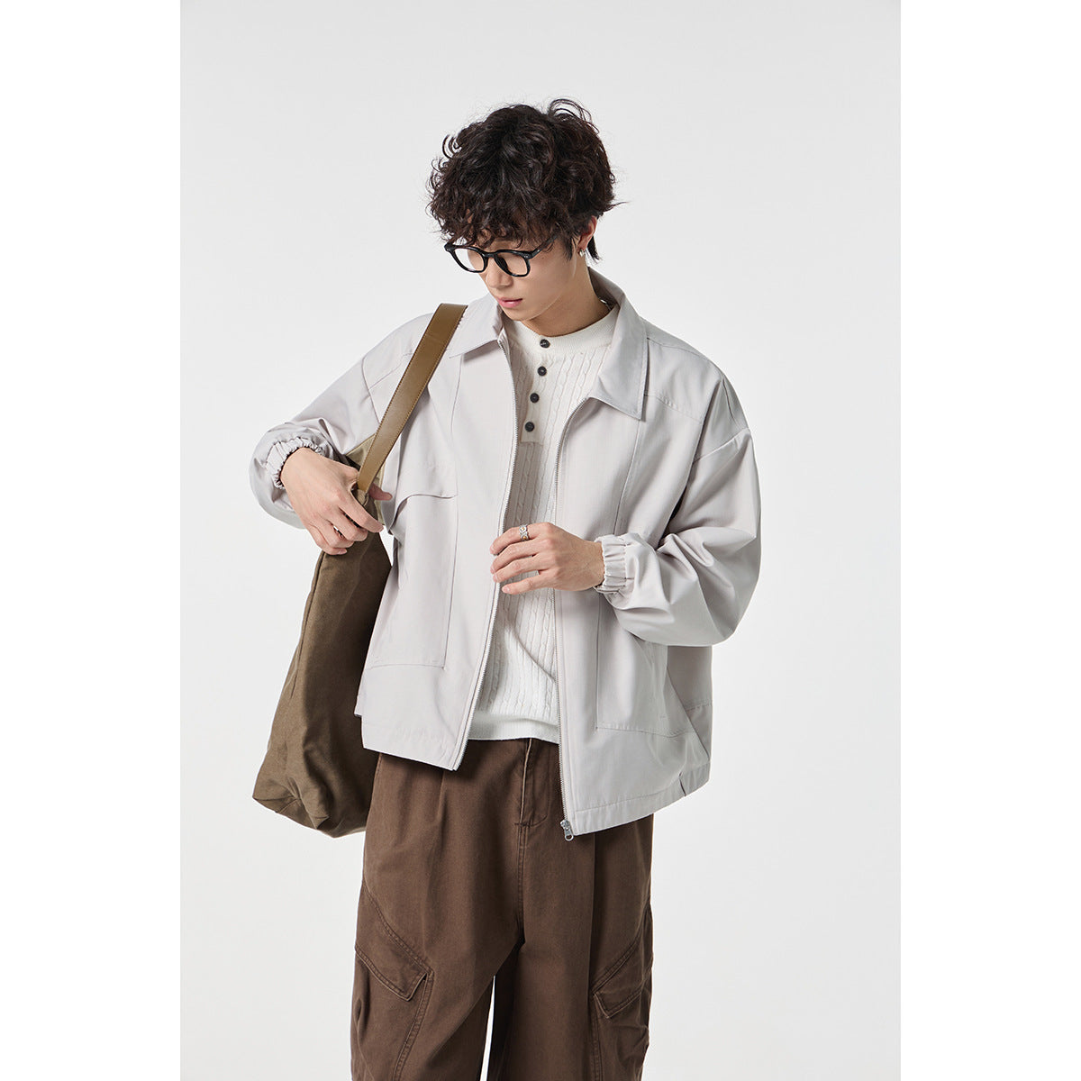 Model wearing a light gray zip-front jacket with a pointed collar and large pockets, styled with brown trousers.